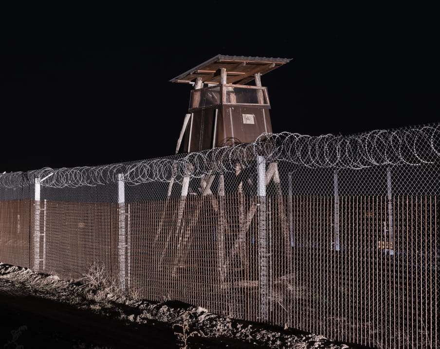 Border barrier topped with electric barbed razor wire and a brown-coloured border guard watchtower on the Hungarian-Serbian border.