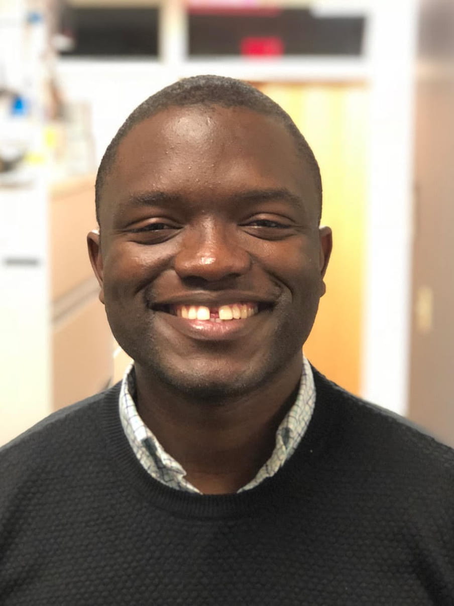 Desmond Jumbam smiling, wearing a collared shirt and a dark grey sweater. 