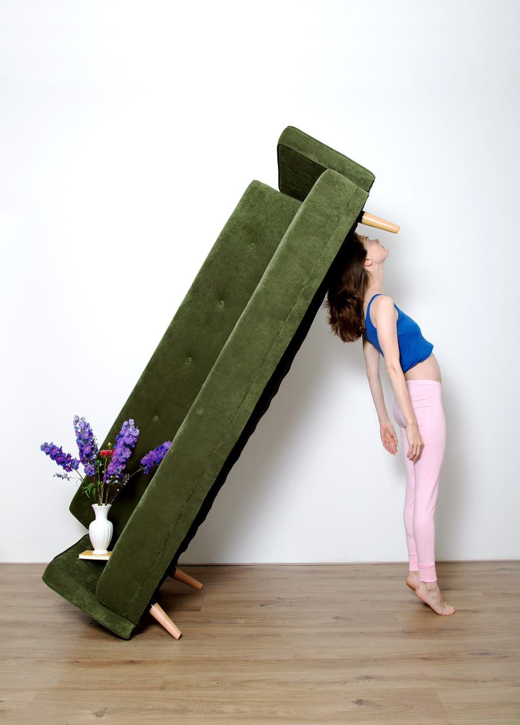 Picture of a female body uplifting a green velvet couch on the side. On the couch there is a flower composition
