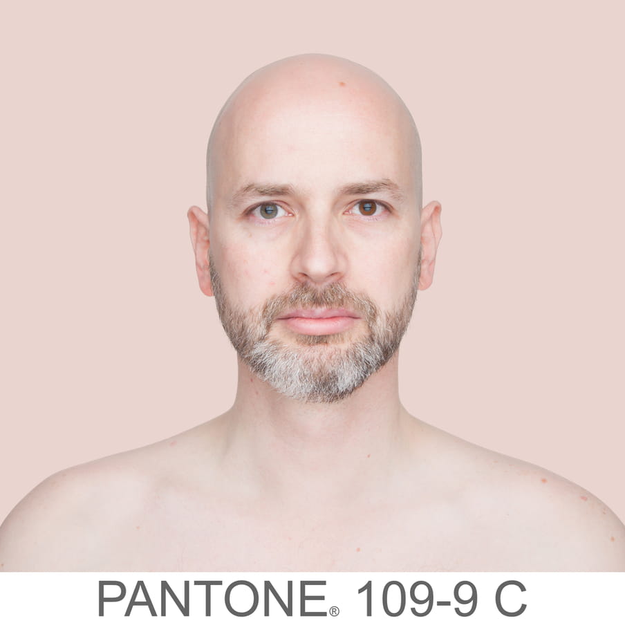 Photographic portrait of a bold man with a beard. The background is a sample of 11 x 11 pixels taken from the nose of the subject and matched with the industrial pallet Pantone®. The pantone color is written out as PANTONE 109-9C. 