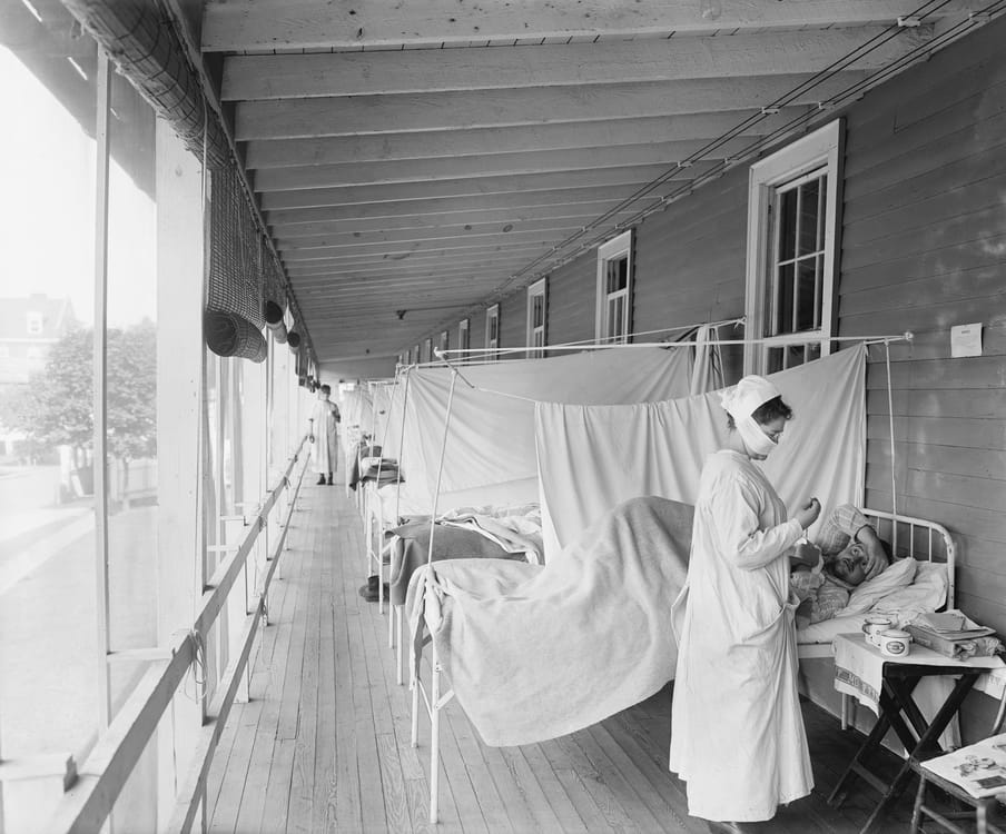 Black and white photograph showing hospital beds aligned on a veranda, separated by sheets. A women wearing a mask is taking care of a man laying in a bed.