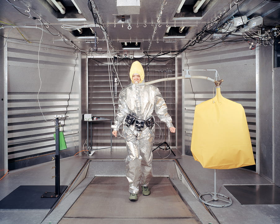 Photo taken in a room with three metallic panels, and silvery light pabels overhead. Walking into a corridor, in a yellow hood and baggy silver space type suit, is a figure with face and hands exposed, green shoes. Lots of slack silvery wires and cables connect them to the ceiling and walls. A yellow inflated sack suspended on a coat rack to the right is connected to her face via an oxygen pump