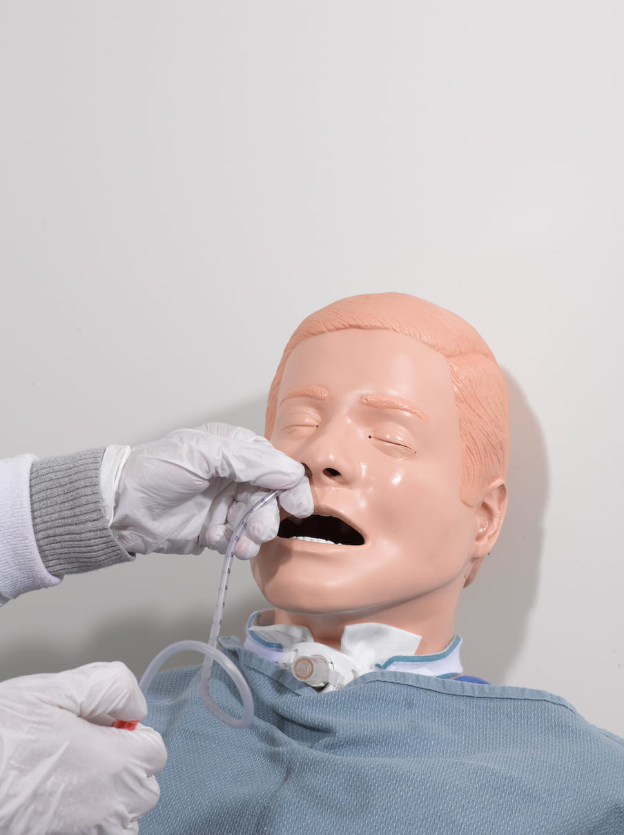 The head of a peach-coloured male mannequin with short hair, eyes closed, is leaned against a white wall, with its mouth open as white gloved hands insert a plastic tube into its left nostril