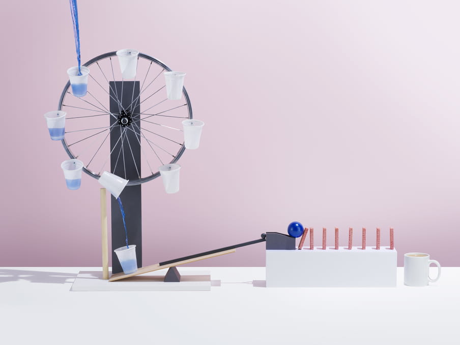 Blue water is poured into one of many rotating white plastic cups attached to a wheel, which pour back down into a cup. This ever filling cup is on a wooden platform connected to a blue ball which rolls onto a stack of pink domino style sticks of sugar. A white mug of brown liquid awaits on the right, for the sugar stick to fall into it. The background is pink.