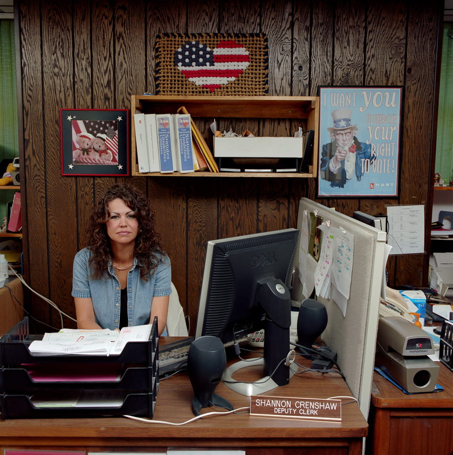 Texan, female deputy clerk sits at a wooden desk. She wears a short-sleeved blouse from jeans fabric. She has long, curly, brown hair. On the desk is a computer, loud speakers, a black filing system and a name plate with her name/job title. We see the edge of another clerk’s desk separated with a cream-coloured partition. The wall behind has some shelves filled with thick guideline books. US voting paraphernalia decorates the wall, including a mounted floor mat showing a heart-shaped US flag.