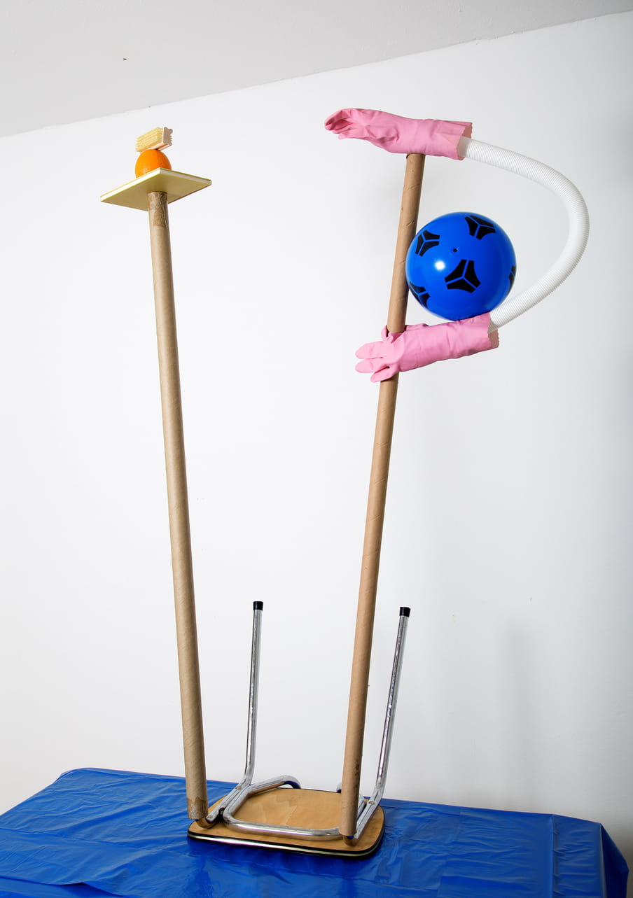 Picture of a house interior with white wall and blue table. On the table a structure composed by a reversed chair with attached two long cardboard tubes. One tube is holding two pink plastic glove and a blue ball and the other one is holding an orange