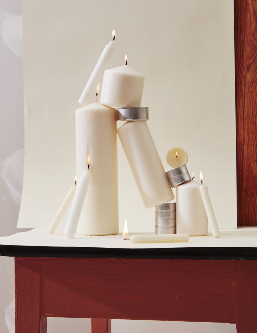 Beige lit candles of various sizes are stack together, in balance, on a brown and black table. The background is also beige with a woody surface visible.