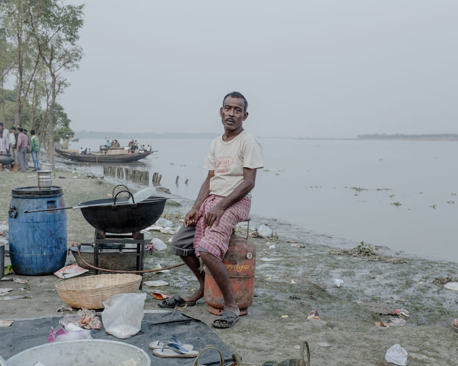 Colour photograph of a man sitting on gaz tank, his back to the water, while looking at the camera. He is wearing a beige t-shirt with something red written on it, grey bermuda pants covered with a piece of pink checked fabric and sandals coming off of his feet. He sits in front a a huge cocking pot. Around him on the muddy ground, are some plastic waste. A group of men are standing in the distance and a boat is floating near the shore.