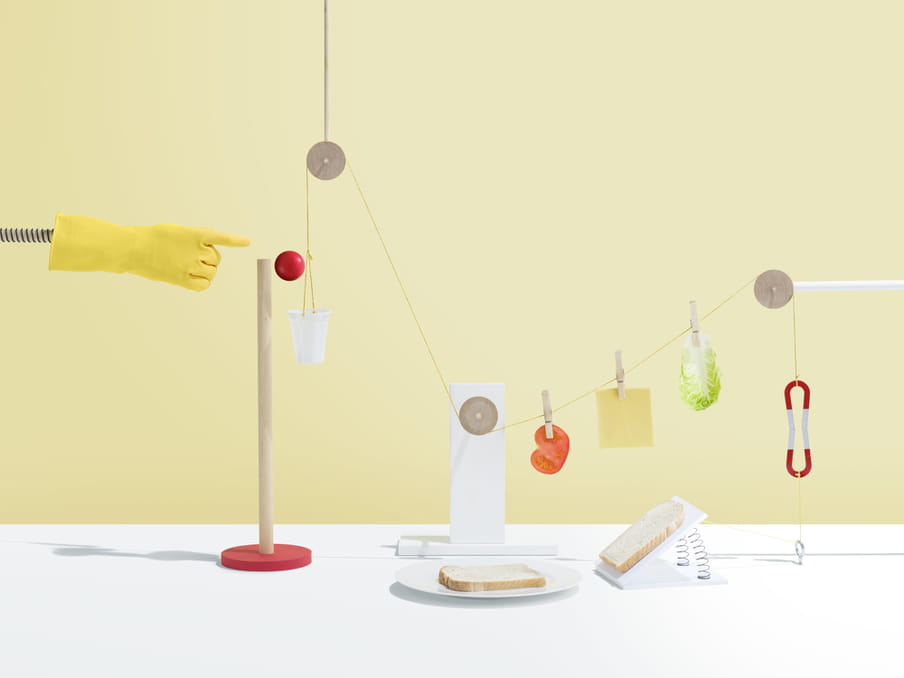 Pieces of salad, cheese and tomato are held onto a string by wooden pegs connected to three circular wooden objects. To the left, a yellow rubber glove on a metal pole seems to have just pushed a red ball off a wooden pole on a stand, and into a white plastic cup. Two pieces of bread lie on a plate and on a stand supported by metal strings.