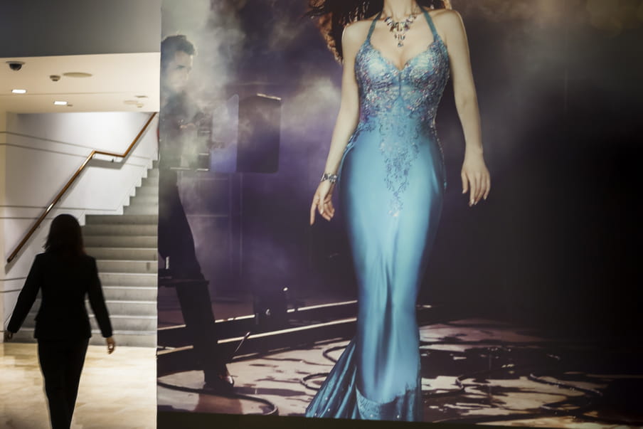 A silhouette of a woman on the left side of the frame walks towards a staircase. On the right is a large poster of a women’s body with a sexy blue dress, lit by a man. 