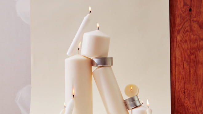 Beige lit candles of various sizes are stack together, in balance. The background is also beige with a woody surface visible. 
