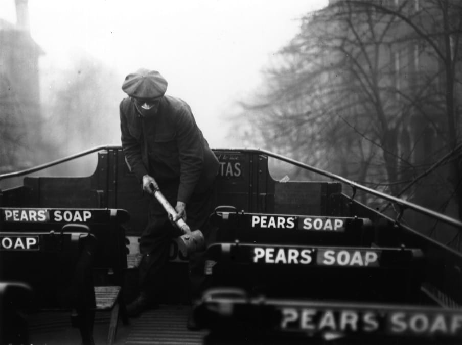 Black and white photograph of a man on the top of the open desk of a bus wearing a mask on his mouth and nose while spaying the seats. 