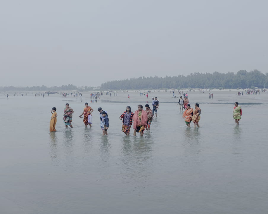 A group of people are walking alone or two by two, holding arms, in the water, pulling their colourful clothes up to their knees. A women is holding her shoes and a bag, a man in the background is carrying a child. We can see the beach and some greenery in the distance with other people standing around.