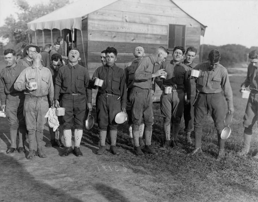 Black and white photograph od a group of men wearing uniforms standing outside and rinsing their mouth. They all hold a cup. Some are smiling, others are spitting. 