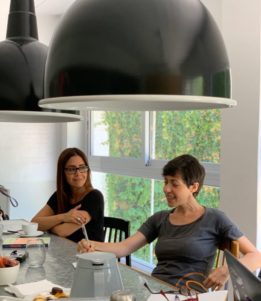 A picture of two women sitting at a table. One to the left is smiling while listening attentively, while another one is talking while writing some notes.