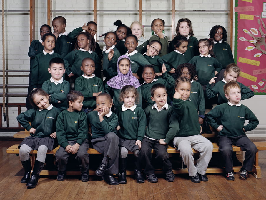 A school photograph of a set of four rows of young happy children posing on benches, with seven at the front, six in the row up, seven in the row behind and eight at the top. They are pulling poses such as with their hands on their hips, posing with chin in hand, pretending to be strong with a fist up at an angle, waving or standing close to another child. The floor is wooden and there is a gymnastic structure behind them, with a children-made poster to the right, in pink. The children wear green jumpers.