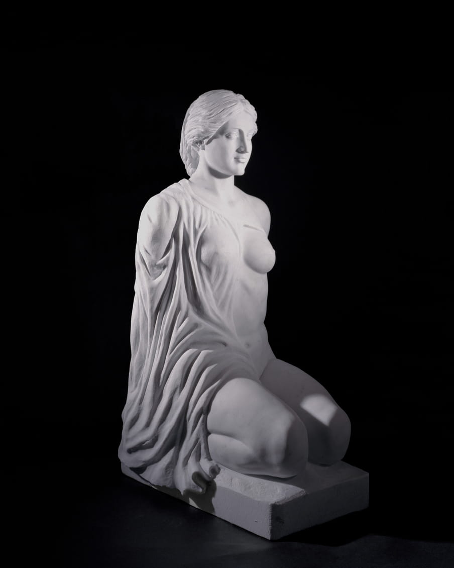 Photo of a plaster statue of a female figure sitting up straight on her knees, her mouth is open, showing teeth, she is wearing a cape, covering one breast - on a black background. 