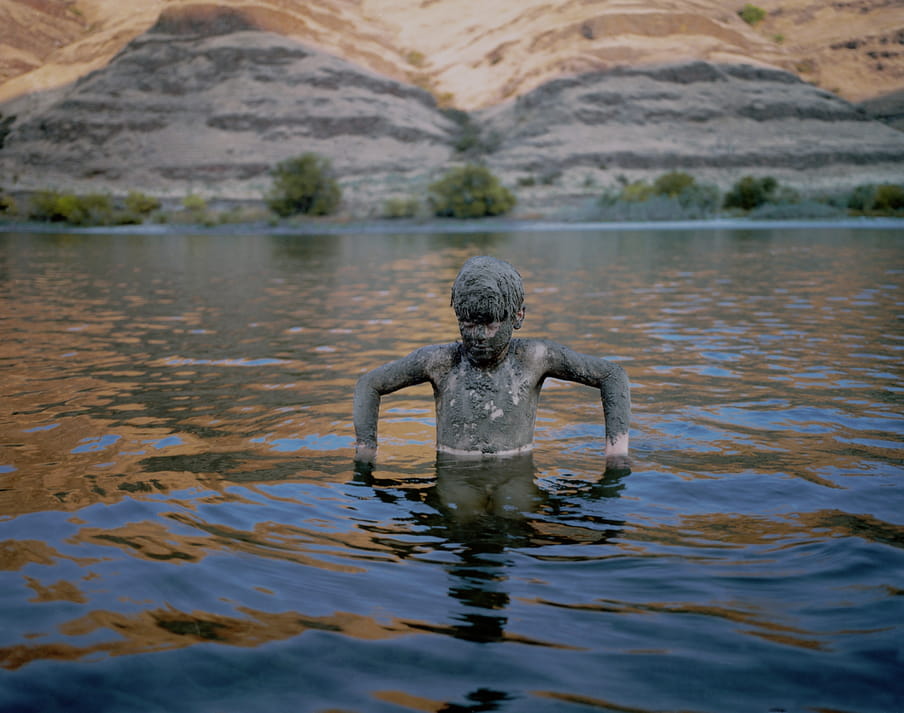 Boy covered in mud, hair and face too, arms out at right angles, standing in a body of water, with sandy mountains behind, the sun hitting their tops, and little grass bushes by the hills.