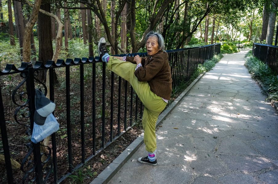 A older person in lime grey jogging pants, a brown top and trainers and pink and blue socks, stretching her leg up high ahainst against a black iron park railing, her hands reaching out to keep the foot up. Their plastic bag with a thermos in and a black and white cap also hang on the railing. The path is lit with sun through the trees