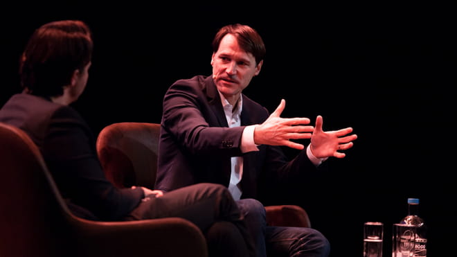 Photo of two men sitting on stage, speaking, one of them gesturing with his hands