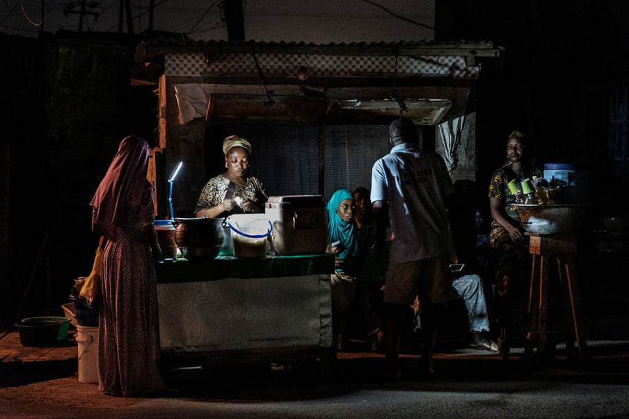 Photograph taken at night with people gathered behind a small stand lit by a precarious lamp. On the left side of the frame, big jars and containers lay on the counter.  