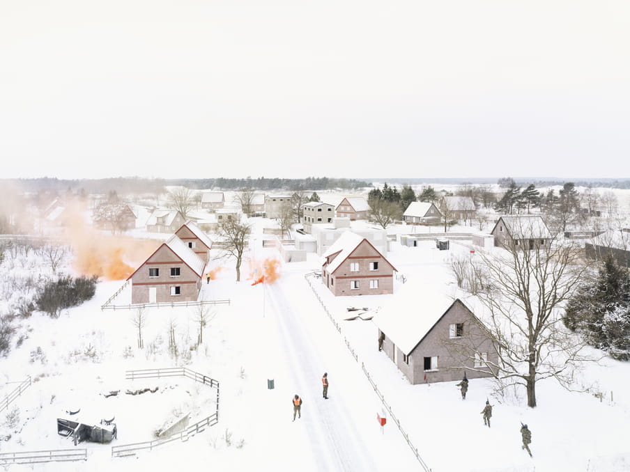 Photo taken from above of a snow-covered village, with three detached houses, a small fenced off circular area, and figures in the distance standing on the road and by a house. The trees are bare, the land looks flat (with more houses and trees in the distance), and there are some orange clouds coming off the ground and next to a house in the distance. The overwhelming feeling is of a white residential landscape