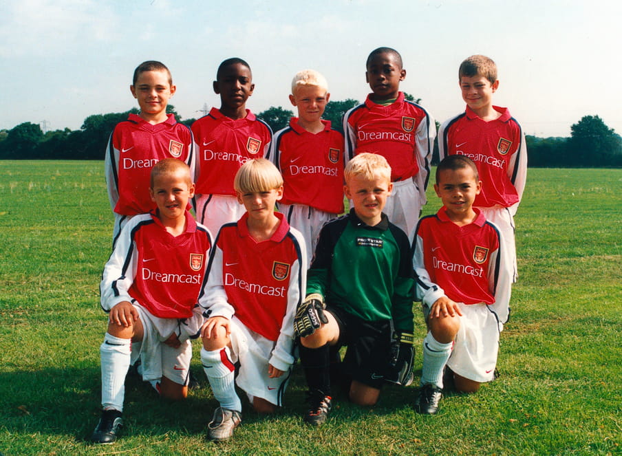 Photo of the Arsenal Advanced team of 2001 taken on a sunny day. A team of 9 boys in total on the green grass of a pitch; 5 standing in the back row and 4 half kneeling in front. Jamie Lawrence is the boy standing in the back right. Harry Kane is bottom left, kneeling.