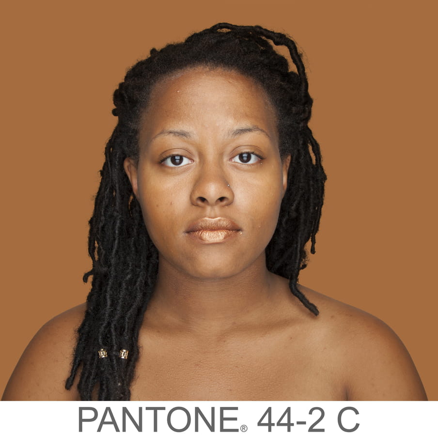 Photographic portrait of a woman with braids and a nose piercing. The background is a sample of 11 x 11 pixels taken from the nose of the subject and matched with the industrial pallet Pantone®. The pantone color is written out as PANTONE 44-2C. 