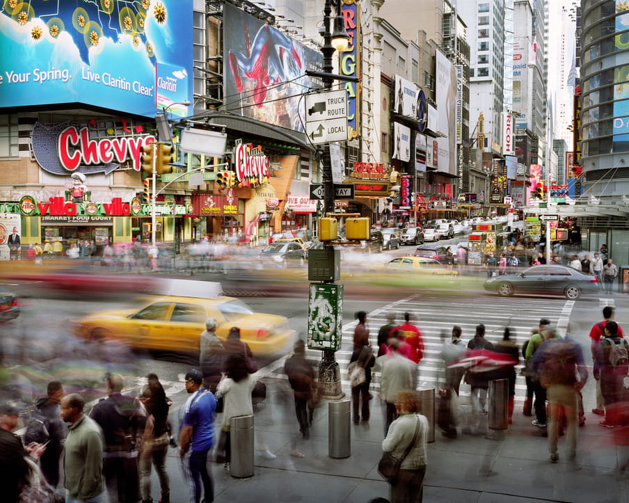 Photo of a busy street with a road trough it, yellow cabs and other cars on it. Many buildings in the back, commercials hanging all over it. It’s shot with a high shutter speed, so people are blurred