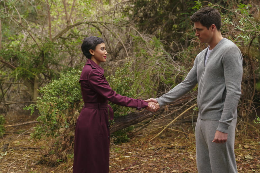 A woman smiling with red lipstick with cropped black hair in a long purple coat shakes hands with a man in grey sweatpants and cardigan, against the backdrop of woods. The handshake is at arm’s length.