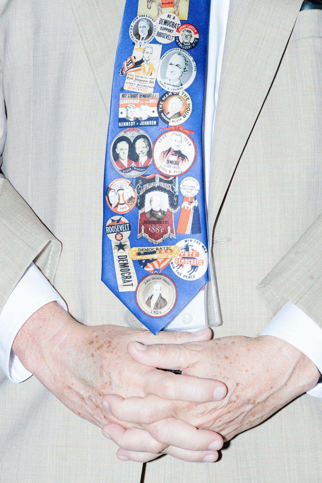 Close-up of folded older looking hands against a pale cream suit, wearing a blue tie covered in political stickers, saying such things as ‘Roosevelt Democrat’ or ‘Kennedy-Johnson’