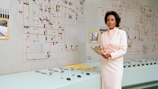 Colour photograph of a woman in a pink suit standing next to a computer.