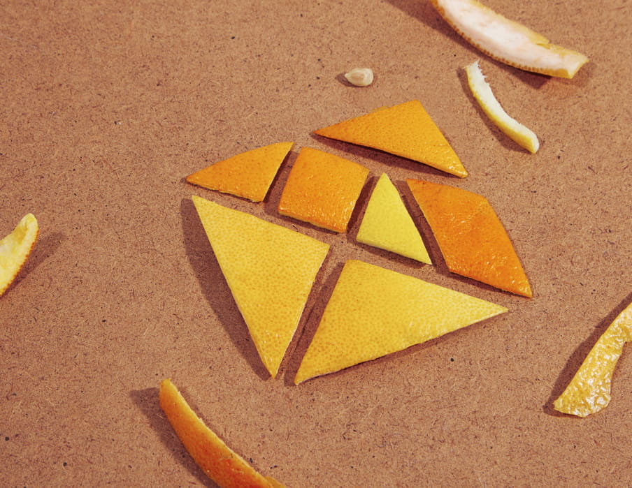 Orange and lemon peels are cut in geometric shapes and arranged to resemble a tangram game. Some extra pieces and a lemon pit lay around the main shape displayed on a woody surface. 