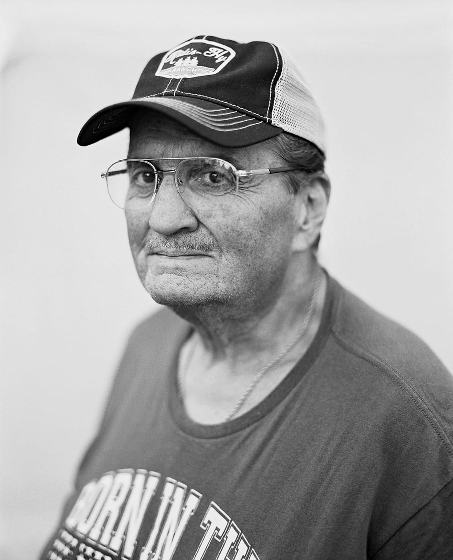 Black and white side headshot portrait of a figure in a black shirt with a cap and beard and glasses, blowing smoke out of his mouth, obscuring his lower face