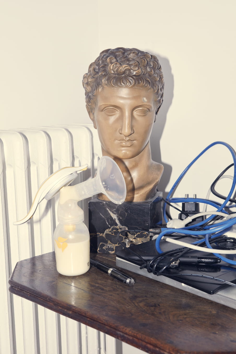 Head of a bronze looking statue on a black marble looking plinth with cables to the right and a breast pump to the left, all on a brown shelf with a white oil radiator to the back