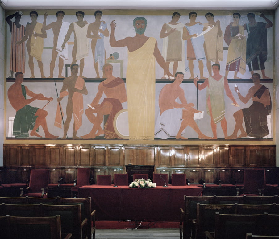 An empty ceremony hall that has been set up for a graduation ceremony. A red velvet cloth covers the table at the top of the room, overlooked by a Greek mural.  