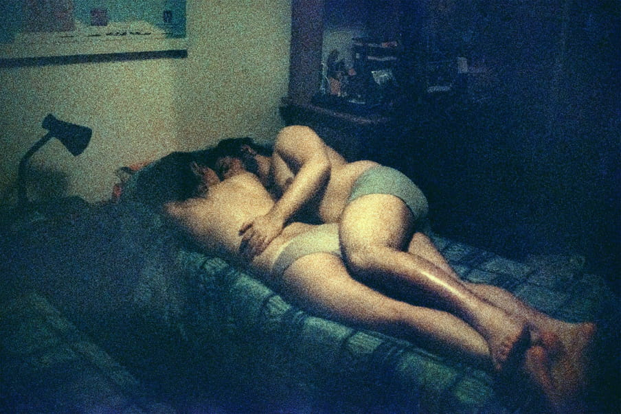 Grainy dark photo of two human figures in white and green underwear hugging on top of the covers of a bed with dark blue and light green patterns. One person lies face down, head turned towards their partner, with black hair, and the partner lies with one leg and arm over their body, faced toward them. Around them is a lamp, a poster on the wall, and a cabinet.