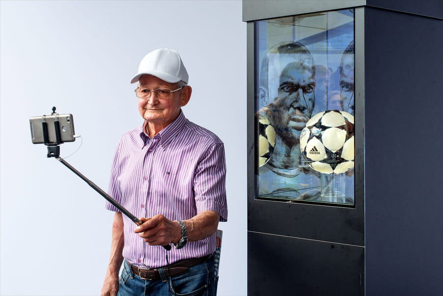 Photo of an older man in a white cap, with glasses, and a striped short-sleeved purple and white shirt tucked into blue jeans with a brown belt and newspaper folded into his back pocket, wearing a watch, holding a selfie stick up as he poses for a self-taken picture with his mobile phone in front of the Champion’s League ball, encased in a glass and black cabinet behind him. There is a black and white drawing of a brooding footballer mirrored on two faces at the back of the cabinet.