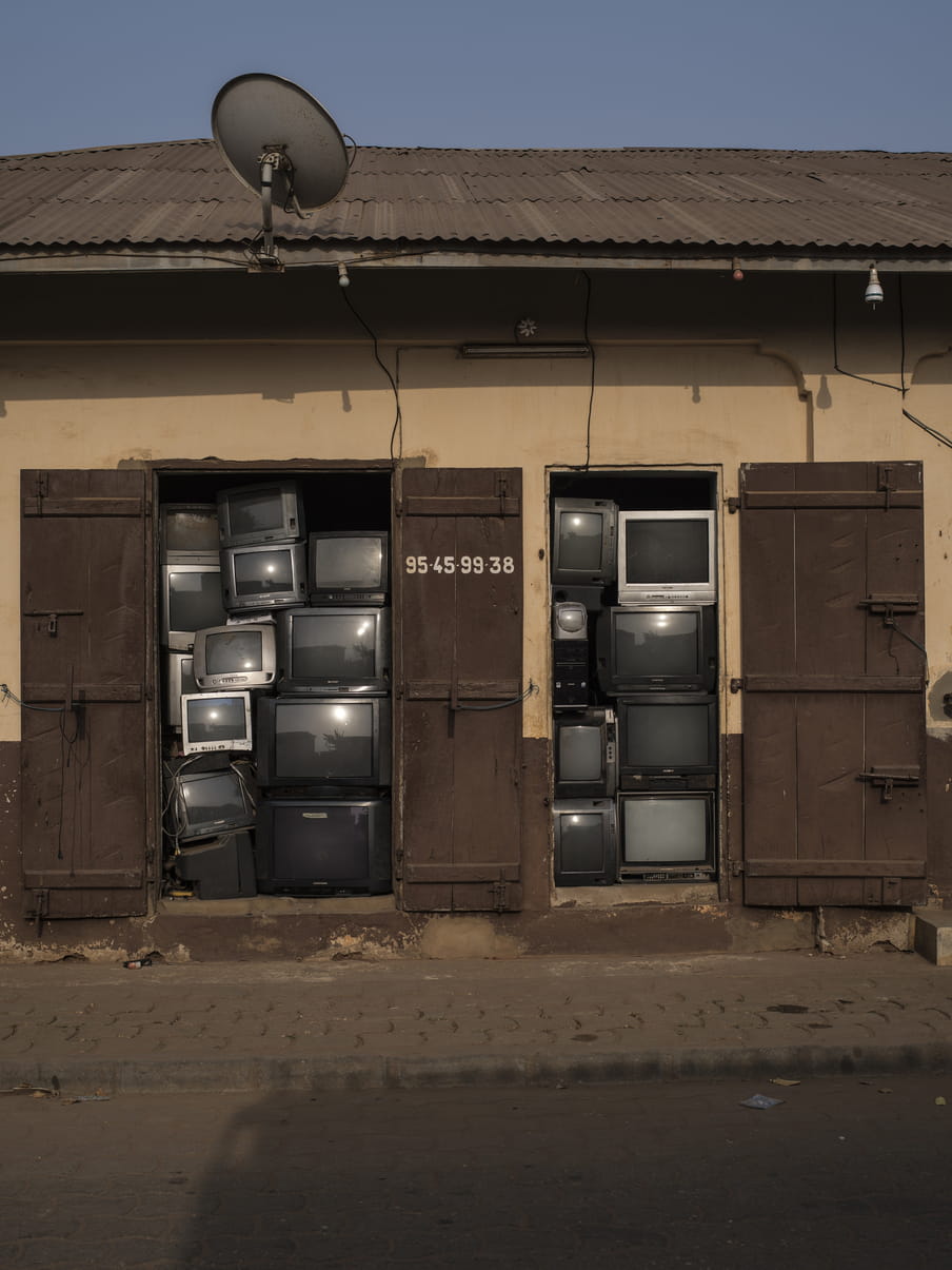 Two large brown wooden sets of doors open on to a narrow pavement, and the doorways are stacked with old televisions; the roof above this building has corrogated iron slates, a satellite dish, and the sky is blue. Photo taken from the middle of the road