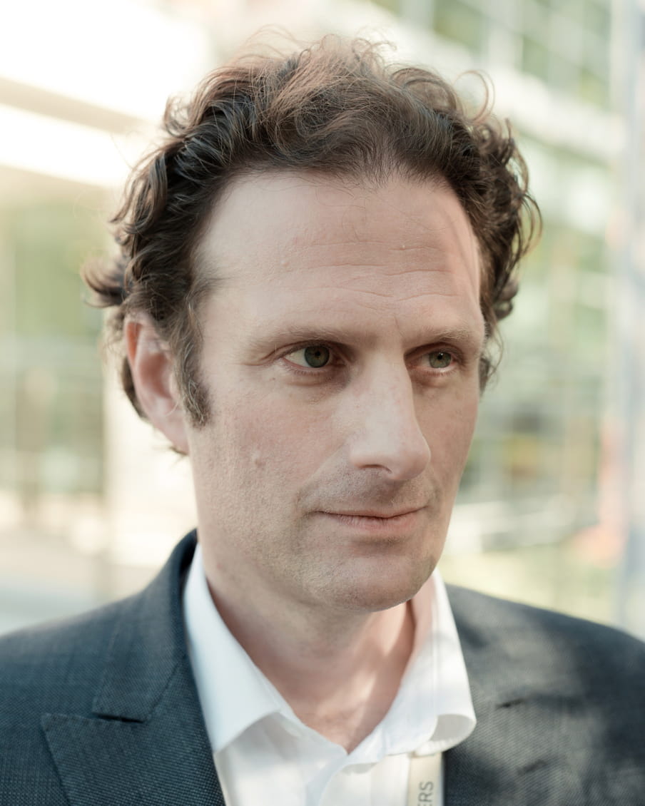 Close up of a mans face, he’s wearing a suit and has short curly hair