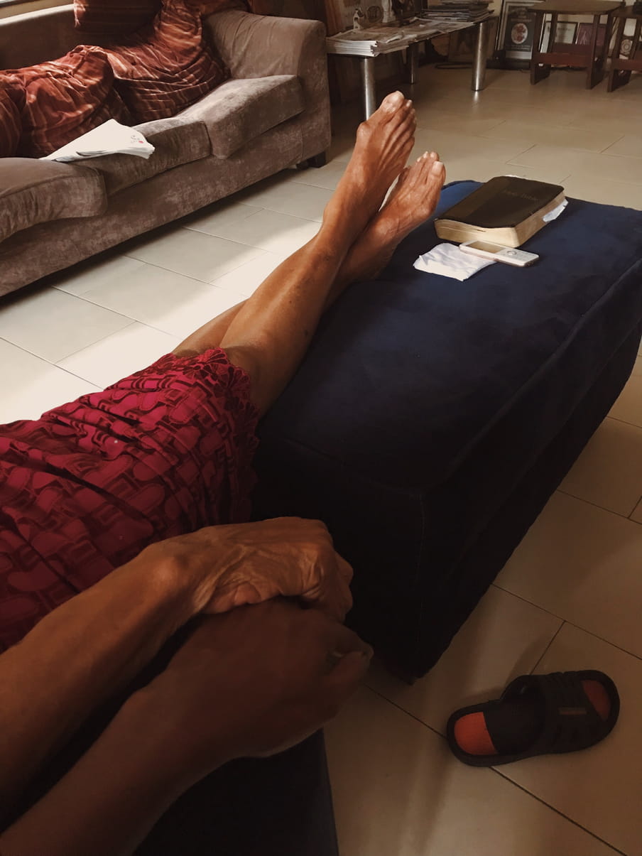 A cropped living room scene in which two dark-skinned women are holding hands. One of them, wearing a maroon textured skirt, is resting both of her legs on a dark blue ottoman. A black book and a white telephone are also placed on the ottoman. 