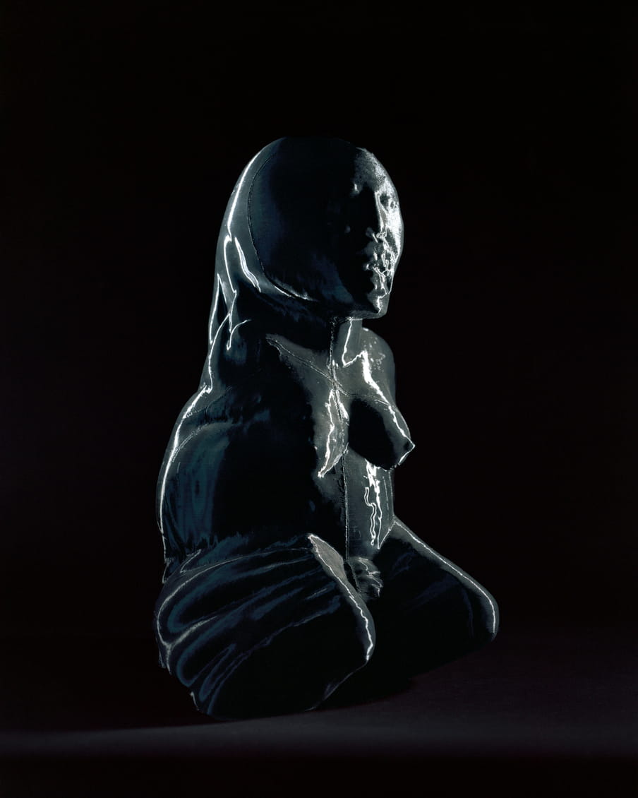 Photo of a plastic looking statue of a female figure giving birth on her knees, her mouth is open, showing teeth - on a black background. Compared to the last image her face is much more distorted and the material is shiny.