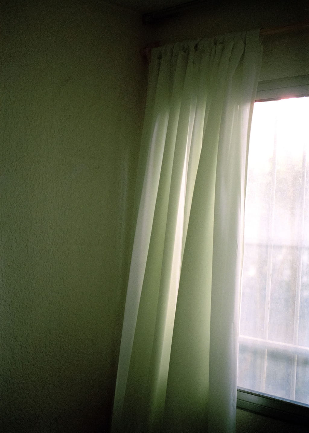 Photo of a closed window with flowing curtains and a wall beside it, with a greenish light around 