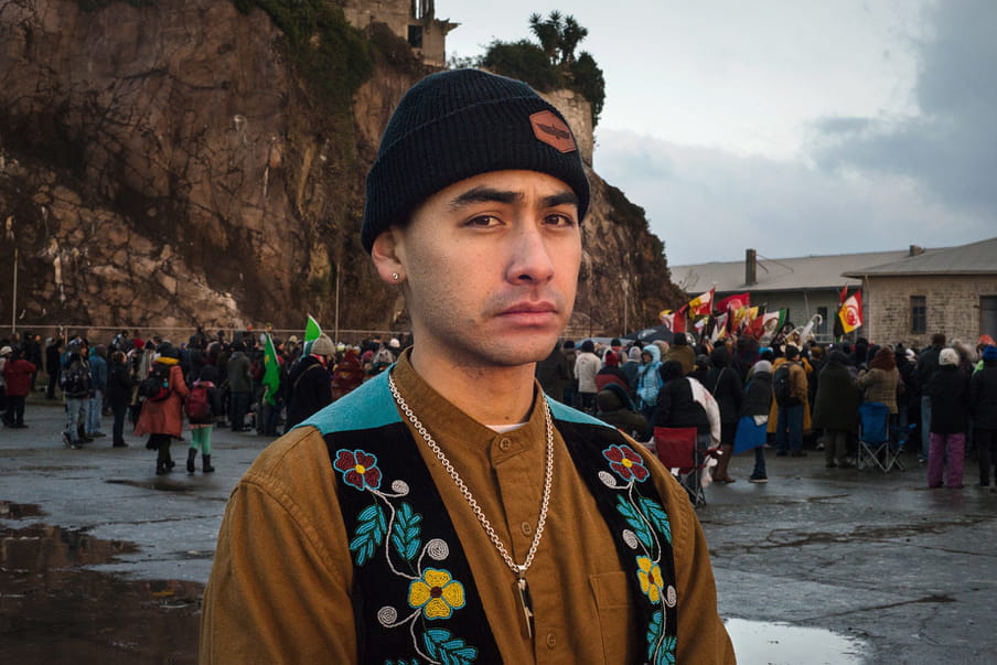 Photo of a young man wearing a orange shirt and an embroidered vest in front of a group of people holding flags