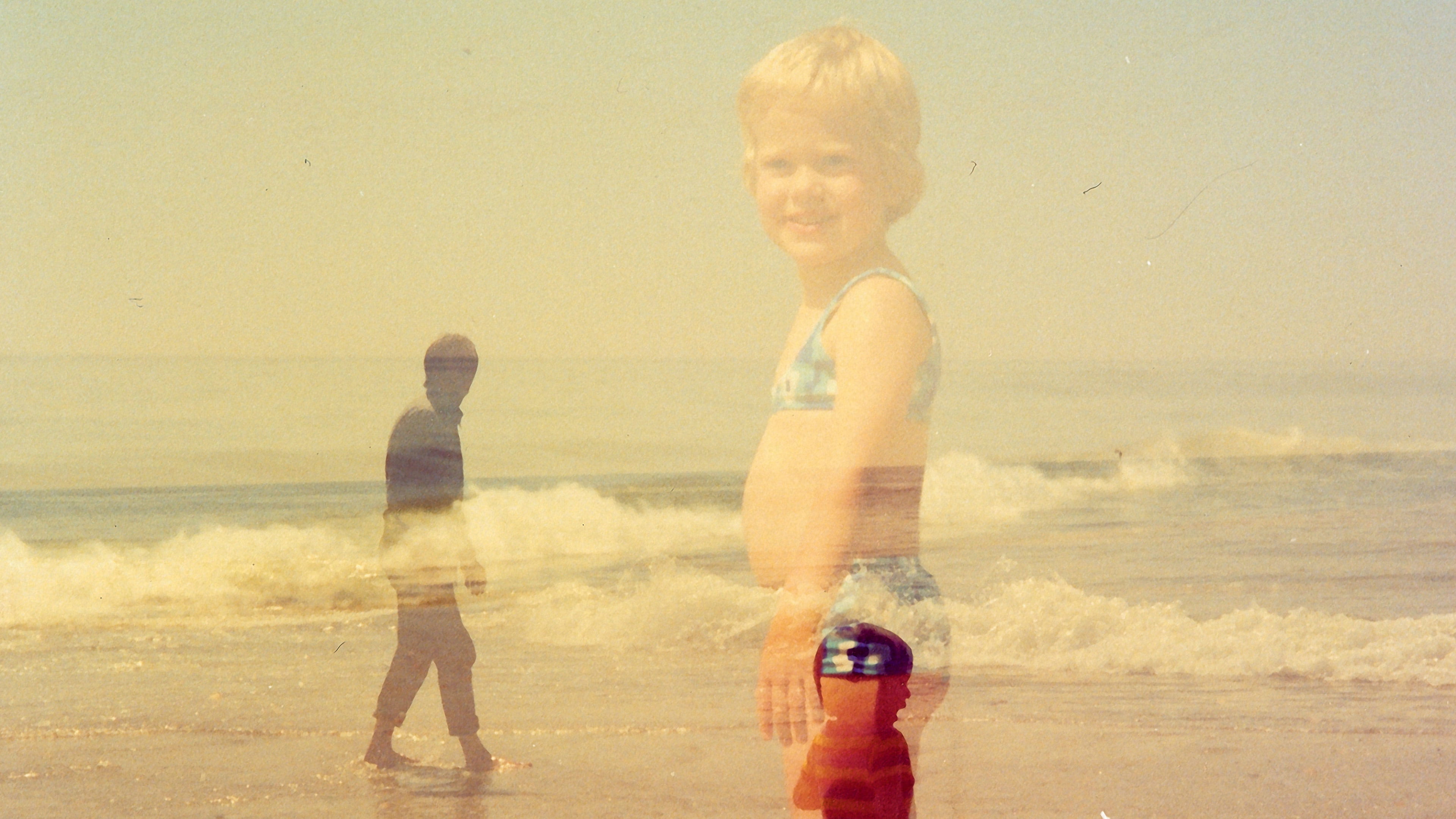 Photo of two kids and a grown man on the beach with waves in the sea on the background. Due to double exposure the opacity of everything is not 100%.