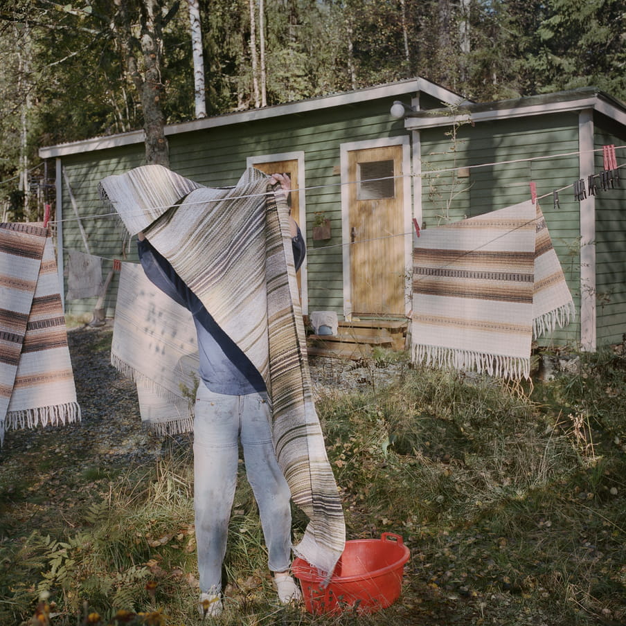 Photograph of a person hanging a rug on a clothesline that is already filled with three other rugs. An orange laundry basket sits besides them in the grass. In the background there is a small green wooden cabin and multiple trees.