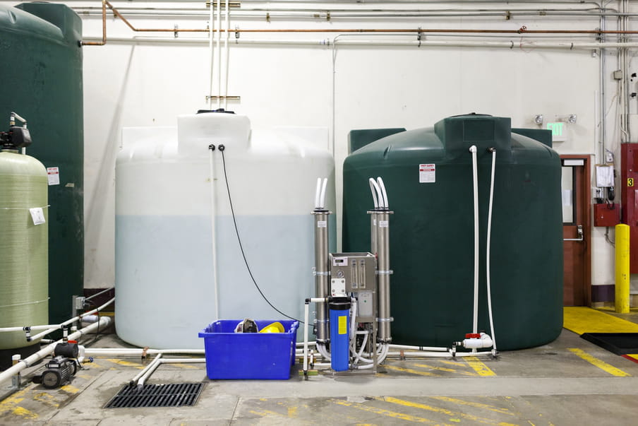 Two big tanks sit side by side on the floor with yellow markings - one is a white almost transprarent tank to the left, and is filled with water, the other is dark green and is filled with nutrients.