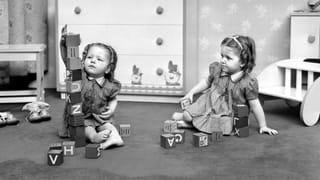Old black and white photo of two little girls sitting on the floor and playing with wooden blocks with letters on them. The left kid has built a high tower out of the blocks, the right one is looking at it while holding a block.