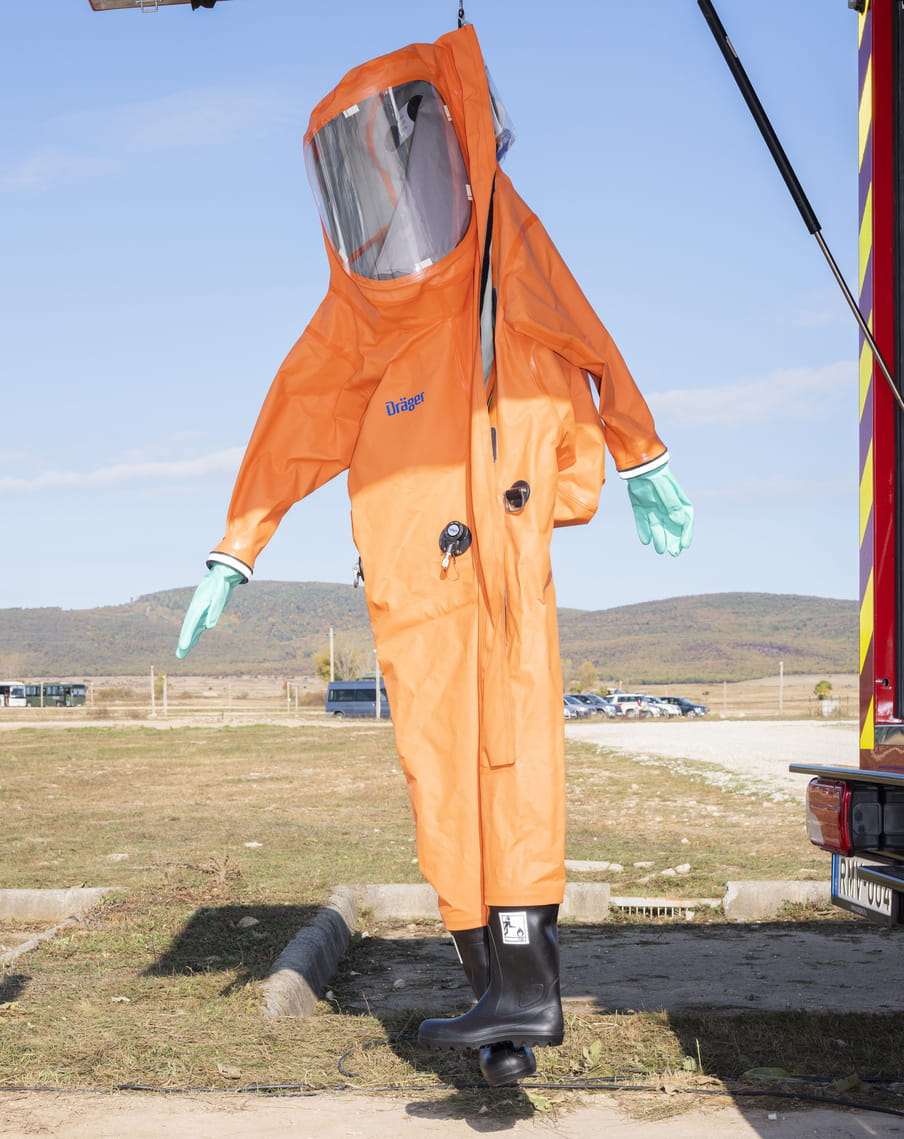 A dummy wearing an orange, chemical protection suit by the brand Dräger with a large, dark, face shield, is fastened to a fire brigade vehicle’s open rear door and dangles from it. The dummy wears teal-coloured gloves and black Wellington boots. It’s part of a military equipment exhibition organised by Hungarian police and military forces. There are forest-lined hills in the background.