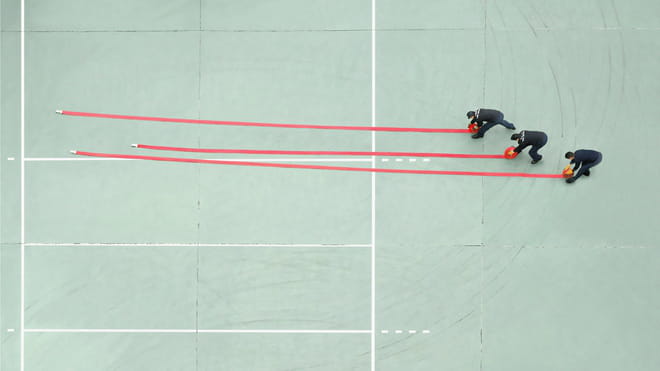 Photograph taken from above of three men unrolling red hoses on a mint green ground court.